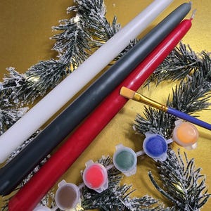 May include: Three plain white, gray, and red candles with a set of six paint pots in various colors, including red, green, blue, orange, brown, and white. The paint pots are arranged in a line on a snowy pine branch.