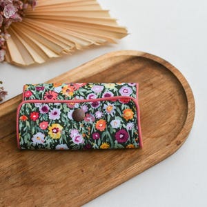 Könnte beinhalten: Ein Portemonnaie mit floralem Muster, rosa Einfassung und Holzknopfverschluss. Das Portemonnaie liegt auf einem Holztablett, im Hintergrund getrocknete Blumen und eine fächerförmige Dekoration. Das Portemonnaie hat ein farbenfrohes Blumenmuster.