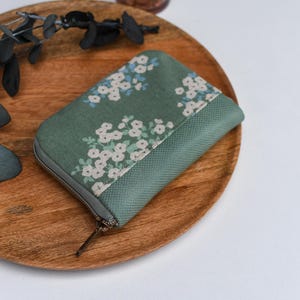 Könnte beinhalten: Eine grüne Reißverschlusstasche mit einem weißen Blumenmuster. Die Tasche liegt auf einer Holzoberfläche.
