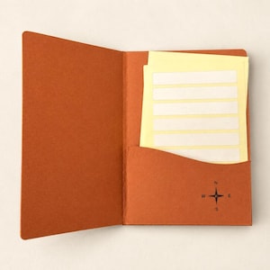 May include: An open rust-colored paper folder with a curved pocket on the right side. The pocket features a small black compass rose icon. Inside the pocket are several stacked pale yellow rectangular labels with white center strips.
