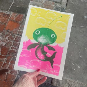 Op de afbeelding: Een kleurrijke print met een grillig wezen met een groene kop, zwarte armen en een roze en zwart lichaam. De achtergrond is verdeeld in gele en roze secties met visachtige vormen.