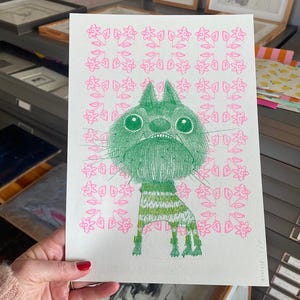 Op de afbeelding: Een grillige illustratie van een groene kat met grote ogen en een chagrijnige uitdrukking. De kat staat tegen een roze bloemenpatroon. De kat draagt een groen-witte gestreepte trui. Het kunstwerk is op wit papier.