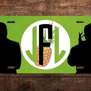 May include: A green and white license plate with a black silhouette of two people on either side. The center of the plate features a white circle with a black letter "F" and a pineapple inside.