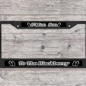 May include: Black license plate frame with white text that reads "C'Mon Son" at the top and "To The Blackberry" at the bottom. The frame has a black background and is set against a wooden background.