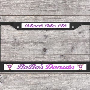 May include: Black license plate frame with the text "Meet Me At" at the top and "BoBo's Donuts" at the bottom in purple. The frame has a cartoon donut graphic on each side of the bottom text.