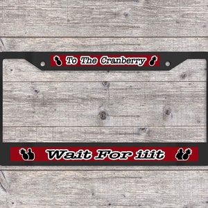 May include: Black metal license plate frame with a red and white stripe across the bottom. The stripe has two black silhouettes of pineapples and the text "Wait For iit". The top stripe is red and has two black silhouettes of pineapples and the text "To The Cranberry".