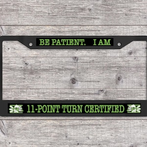 May include: Black license plate frame with the text "BE PATIENT. I AM" at the top and "11-POINT TURN CERTIFIED" at the bottom, in green lettering. The frame has a rectangular shape and is set against a wooden background.