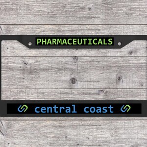 May include: Black license plate frame with the text "PHARMACEUTICALS" in green and "central coast" in blue. The frame has a rectangular shape with rounded corners and is made of a matte black material.