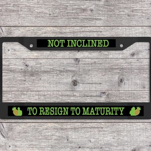 May include: Black license plate frame with the text "NOT INCLINED" and "TO RESIGN TO MATURITY" in green. The frame has a rectangular shape with rounded corners and a black background. Pineapple graphics are on each side.