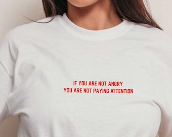 May include: White t-shirt with red text that reads "If you are not angry you are not paying attention."