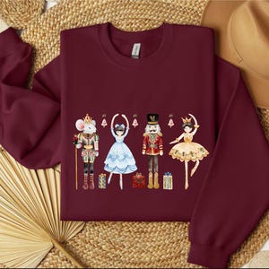 Könnte beinhalten: Burgunderfarbenes Sweatshirt mit Rundhalsausschnitt und einem Nussknacker-Motiv. Die Grafik zeigt eine Maus, eine Ballerina, einen Nussknackersoldaten und eine Ballerina, alle in festlicher Kleidung. Geschenke und Ornamente sind ebenfalls im Design enthalten.