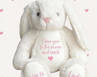 Op de afbeelding: Een witte pluche konijn met lange oren, een roze neus en een hartvormig ontwerp. De tekst "I love you to the moon and back" staat op de borst van het konijn, samen met een roze hart. De datum "14 feb 26" en de naam "Linda" staan op de voeten van het konijn.
