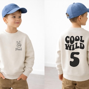 Könnte beinhalten: Beiges Sweatshirt mit schwarzen Grafiken. Auf der Vorderseite befindet sich ein Friedenszeichen und der Name "Mark". Auf der Rückseite steht "COOL WILD 5". Das Kind trägt eine blaue Baseballkappe und eine braune Hose. Ein lässiges Outfit für einen Geburtstag.