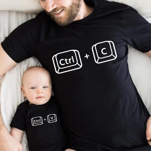 May include: Black t-shirt with white "Ctrl + C" graphic. A baby in a black onesie with a white "Ctrl + V" graphic is next to the shirt. The image shows a parent and child wearing matching outfits, a fun and playful theme.