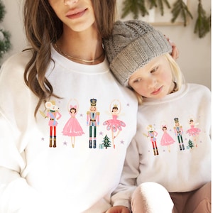 Könnte beinhalten: Weiße Sweatshirts mit einem Nussknacker-Motiv. Das Design zeigt eine Maus, eine Ballerina, einen Nussknacker-Soldaten und einen Weihnachtsbaum. Die Sweatshirts werden von zwei Personen getragen.
