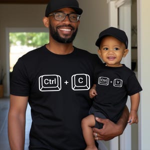 May include: Black t-shirts with white keyboard command graphics. One shirt displays "Ctrl + C", the other "Ctrl + V". The shirts are worn by a father and child, both wearing black caps and glasses.