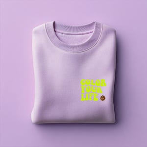 Könnte beinhalten: Ein gefalteter, fliederfarbener Pullover mit dem Text "COLOR YOUR LIFE" in leuchtendem Gelb und einem kleinen Leopardenmuster-Kreis. Der Pullover liegt auf einer passenden fliederfarbenen Oberfläche.