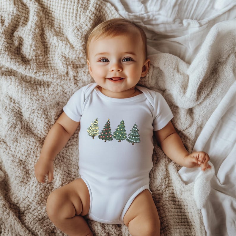 May include: A white baby onesie featuring a row of colorful Christmas tree designs. The trees are in shades of green, gold, and red, with various decorative elements. The baby is lying on a soft, textured blanket.