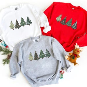 May include: Three sweatshirts in white, red, and gray, each featuring a row of embroidered Christmas trees. The trees vary in design, with some adorned with lights and ornaments. A festive holiday theme.