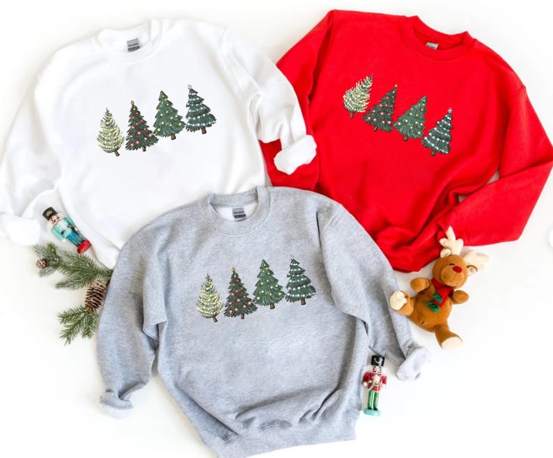 May include: Three sweatshirts in white, red, and gray, each featuring a row of embroidered Christmas trees. The trees vary in design, with some adorned with lights and ornaments. A festive holiday theme.
