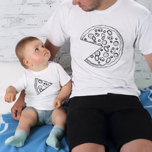 以下が含まれることがあります： ピザのスライスデザインが施されたお揃いの白いTシャツ。1枚のシャツには、欠けているスライスのあるピザ全体が描かれ、もう1枚には1つのスライスが描かれています。シャツは親と子供が着用し、楽しくコーディネートされた外観を作り出しています。