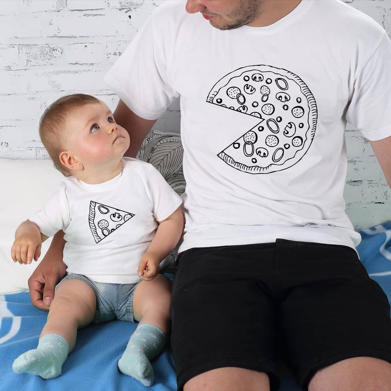 以下が含まれることがあります： ピザのスライスデザインが施されたお揃いの白いTシャツ。1枚のシャツには、欠けているスライスのあるピザ全体が描かれ、もう1枚には1つのスライスが描かれています。シャツは親と子供が着用し、楽しくコーディネートされた外観を作り出しています。