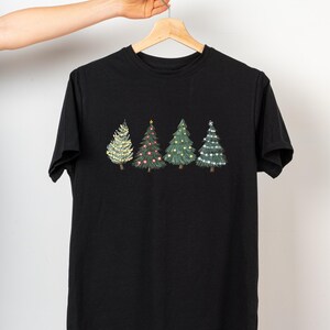 May include: Black t-shirt featuring four embroidered Christmas trees. The trees are decorated with lights and ornaments in various colors, including white, red, and gold. The trees are arranged in a row across the chest of the shirt.