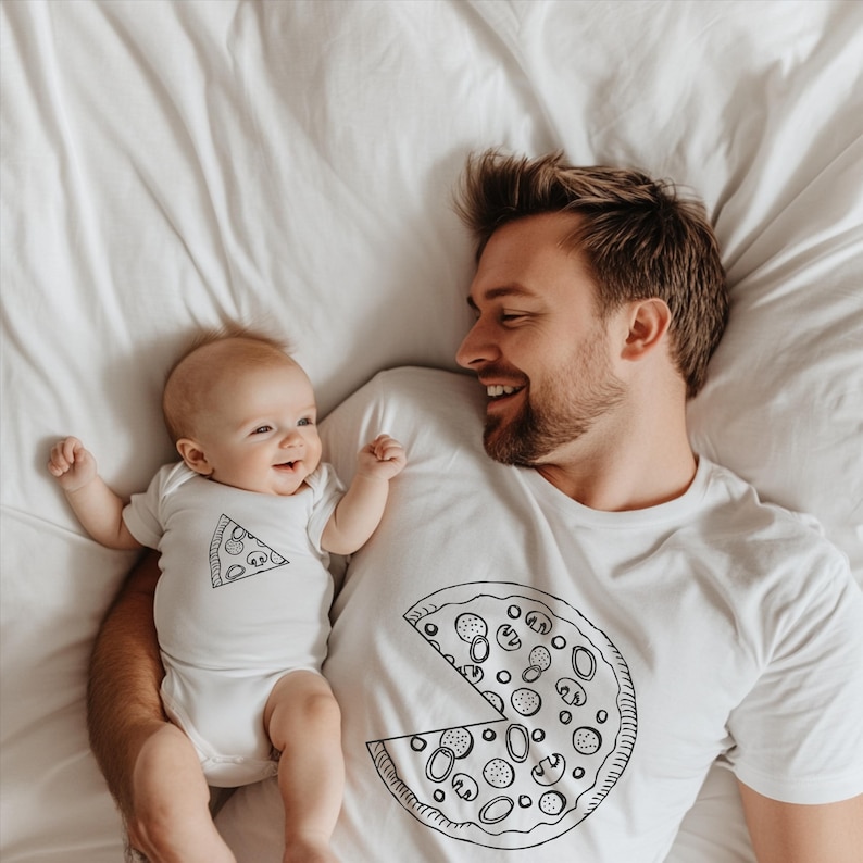 以下が含まれることがあります： ピザのスライスグラフィックの白いボディスーツと、ピザグラフィックの白いTシャツ。赤ちゃんとおとなは白いシーツの上にいます。大人は笑顔です。