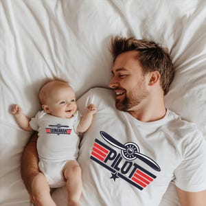 Könnte beinhalten: Ein Baby in einem weißen Body mit dem Text "WINGMAN" in Rot und Blau, und ein Erwachsener in einem weißen T-Shirt mit einem Propeller-Grafik und dem Wort "PILOT" in Rot und Blau. Beide liegen auf einem weißen Bettlaken.