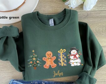 May include: Bottle green Christmas sweatshirt with a festive design. The design includes a Christmas tree, gingerbread man, wrapped gift, and snowman. The name "John" is printed below the design.