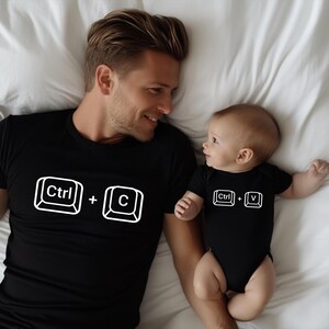 May include: Black t-shirt with a white "Ctrl + C" graphic and a black baby onesie with a "Ctrl + V" graphic. The shirt and onesie are worn by a man and a baby, respectively, lying on a white bedsheet.