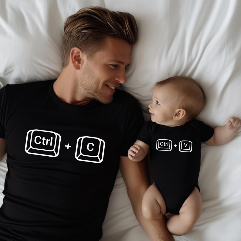 May include: Black t-shirt with a white "Ctrl + C" graphic and a black baby onesie with a "Ctrl + V" graphic. The shirt and onesie are worn by a man and a baby, respectively, lying on a white bedsheet.