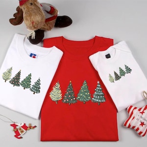 May include: Three Christmas-themed t-shirts and a onesie. The shirts are white and red, and the onesie is white. Each garment features a row of decorated Christmas trees in green, gold, and red. A plush reindeer and ornaments are also in the image.