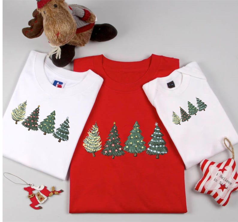 May include: Three Christmas-themed t-shirts and a onesie. The shirts are white and red, and the onesie is white. Each garment features a row of decorated Christmas trees in green, gold, and red. A plush reindeer and ornaments are also in the image.