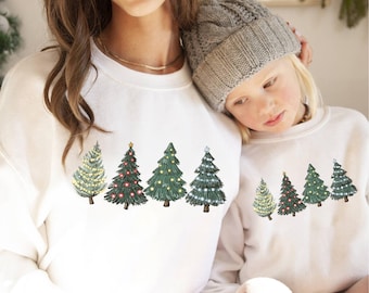 Könnte beinhalten: Weiße Sweatshirts mit einer Reihe von Weihnachtsbäumen, die mit Lichtern und Sternen verziert sind. Die Bäume sind in Grüntönen gehalten, mit roten und weißen Lichtern. Getragen von zwei Personen.