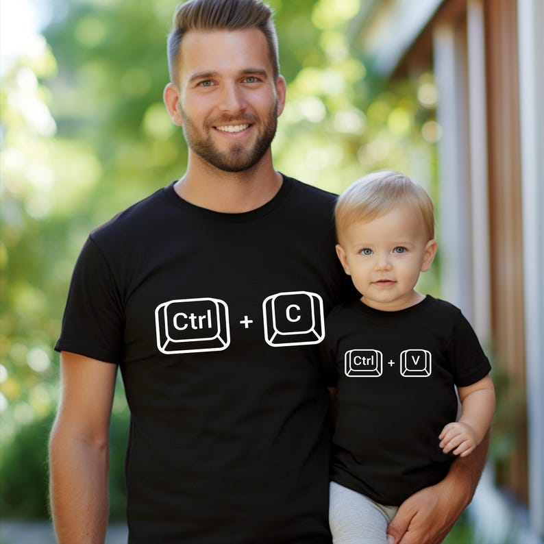 May include: Matching black t-shirts with white text. One shirt has "Ctrl + C" and the other has "Ctrl + V". The shirts are worn by a man and a baby. The image is taken outdoors.
