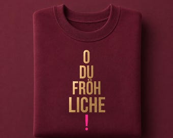 May include: A burgundy crewneck sweatshirt featuring gold metallic text arranged in a triangular shape that reads O DU FRÖHLICHE with a bright pink exclamation point at the bottom. The garment has a ribbed collar and long sleeves.