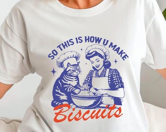Può includere: Maglietta bianca con un'illustrazione blu di un gatto con un cappello da cuoco e una donna con un cappello da cuoco che fa biscotti. Il testo sulla maglietta recita "So this is how u make biscuits".