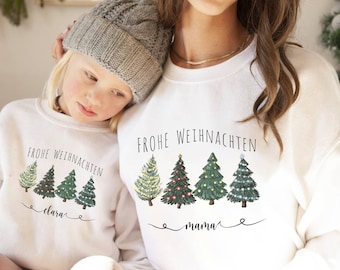 Könnte beinhalten: Zwei weiße Sweatshirts mit Rundhalsausschnitt und dem Text "FROHE WEIHNACHTEN" über einer Reihe von vier illustrierten Weihnachtsbäumen. Das kleinere Sweatshirt trägt den Namen "clara" und das größere den Namen "mama".
