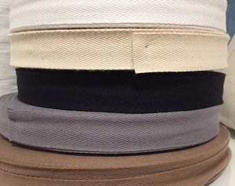 May include: Five spools of cotton twill tape in different colours. The spools are stacked on top of each other. The colours are white, beige, black, grey, and brown.