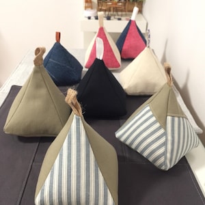 May include: A set of eight triangular doorstops in various fabrics, including denim, striped, and solid colours. The doorstops are arranged on a dark grey surface.