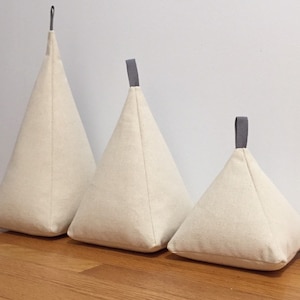 May include: Three beige fabric doorstops in the shape of a pyramid with grey fabric handles. The doorstops are arranged in order of size from largest to smallest.