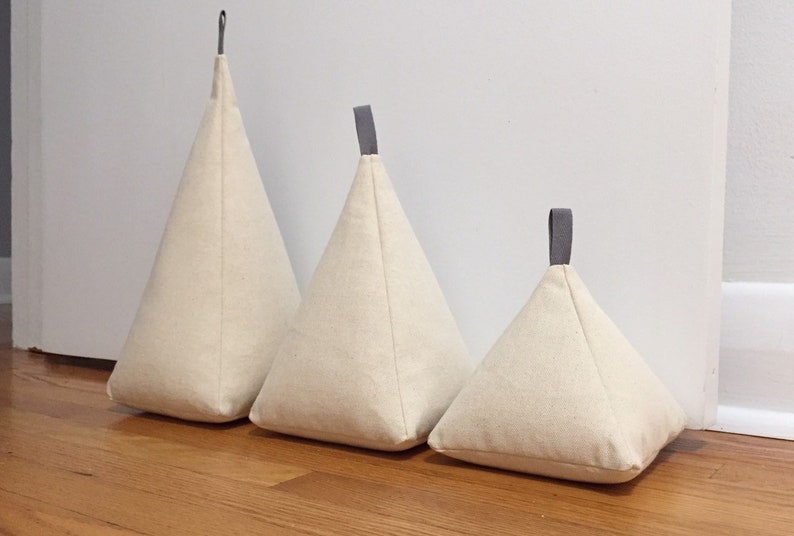 May include: Three beige fabric doorstops in the shape of a pyramid with grey fabric handles. The doorstops are arranged in order of size from largest to smallest.
