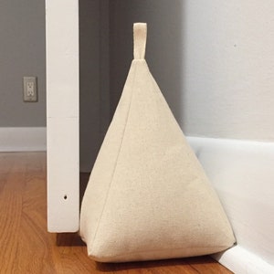 May include: A beige fabric doorstop shaped like a pyramid with a loop handle. The doorstop is positioned against a white door frame and a white skirting board.