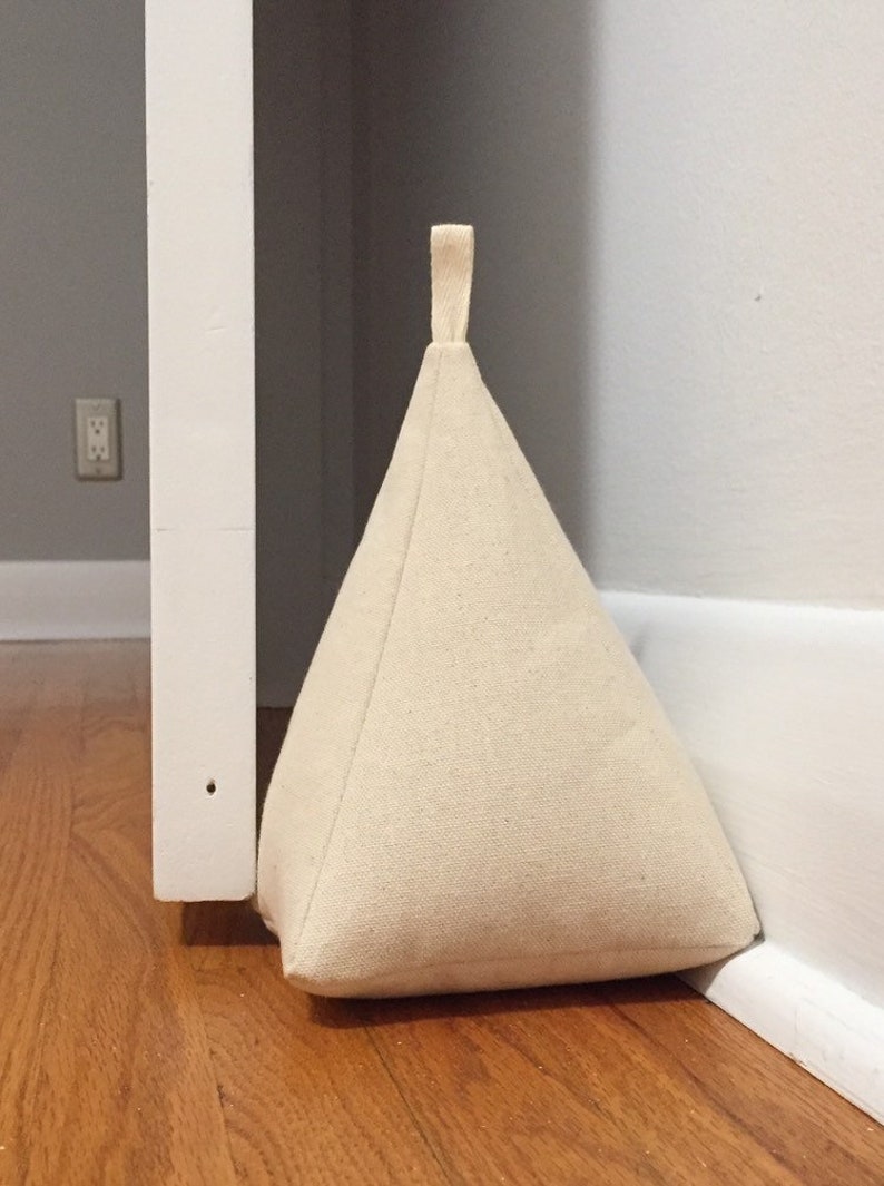 May include: A beige fabric doorstop shaped like a pyramid with a loop handle. The doorstop is positioned against a white door frame and a white skirting board.