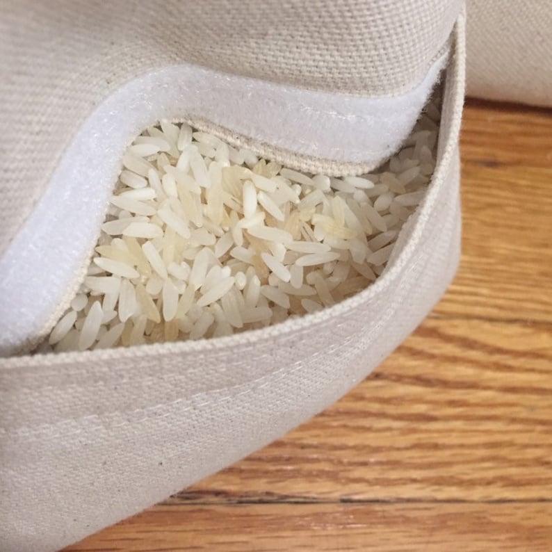 May include: A close-up of a white canvas bag filled with white rice. The bag has a white zipper and is sitting on a wooden surface.