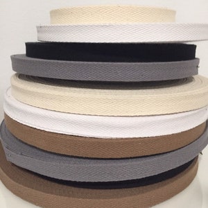 May include: A stack of different coloured cotton twill tape, also known as herringbone tape. The tape is woven with a chevron pattern and is available in beige, white, black, brown, and grey.