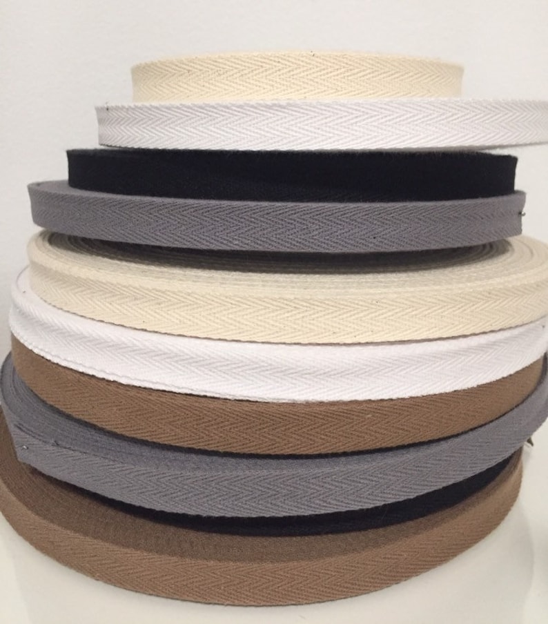 May include: A stack of different coloured cotton twill tape, also known as herringbone tape. The tape is woven with a chevron pattern and is available in beige, white, black, brown, and grey.