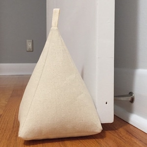 May include: A beige fabric doorstop in the shape of a triangle with a loop handle. The doorstop is wedged against a white door.