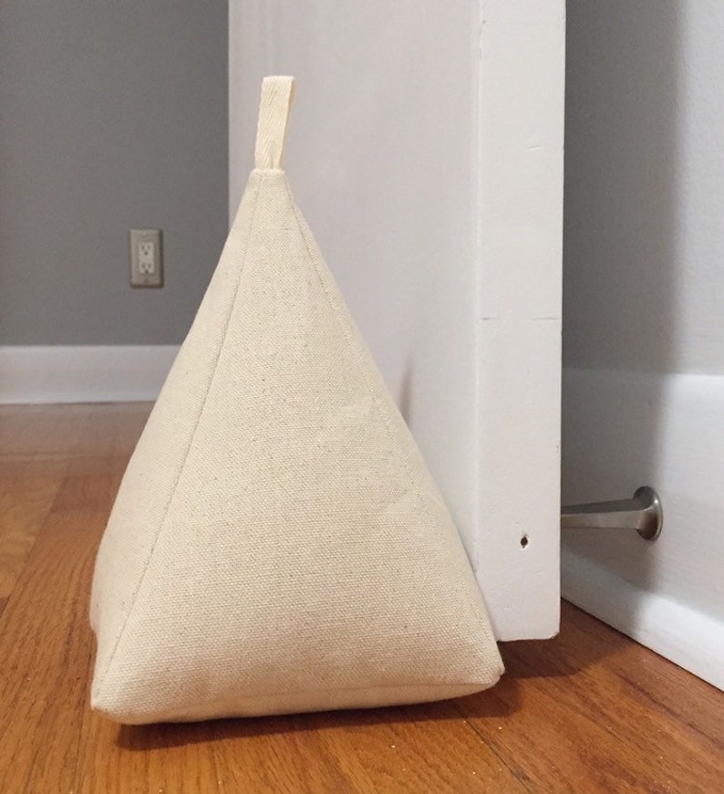 May include: A beige fabric doorstop in the shape of a triangle with a loop handle. The doorstop is wedged against a white door.